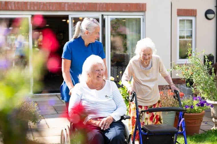 A UK care home carer supporting an elderly resident in a communal lounge